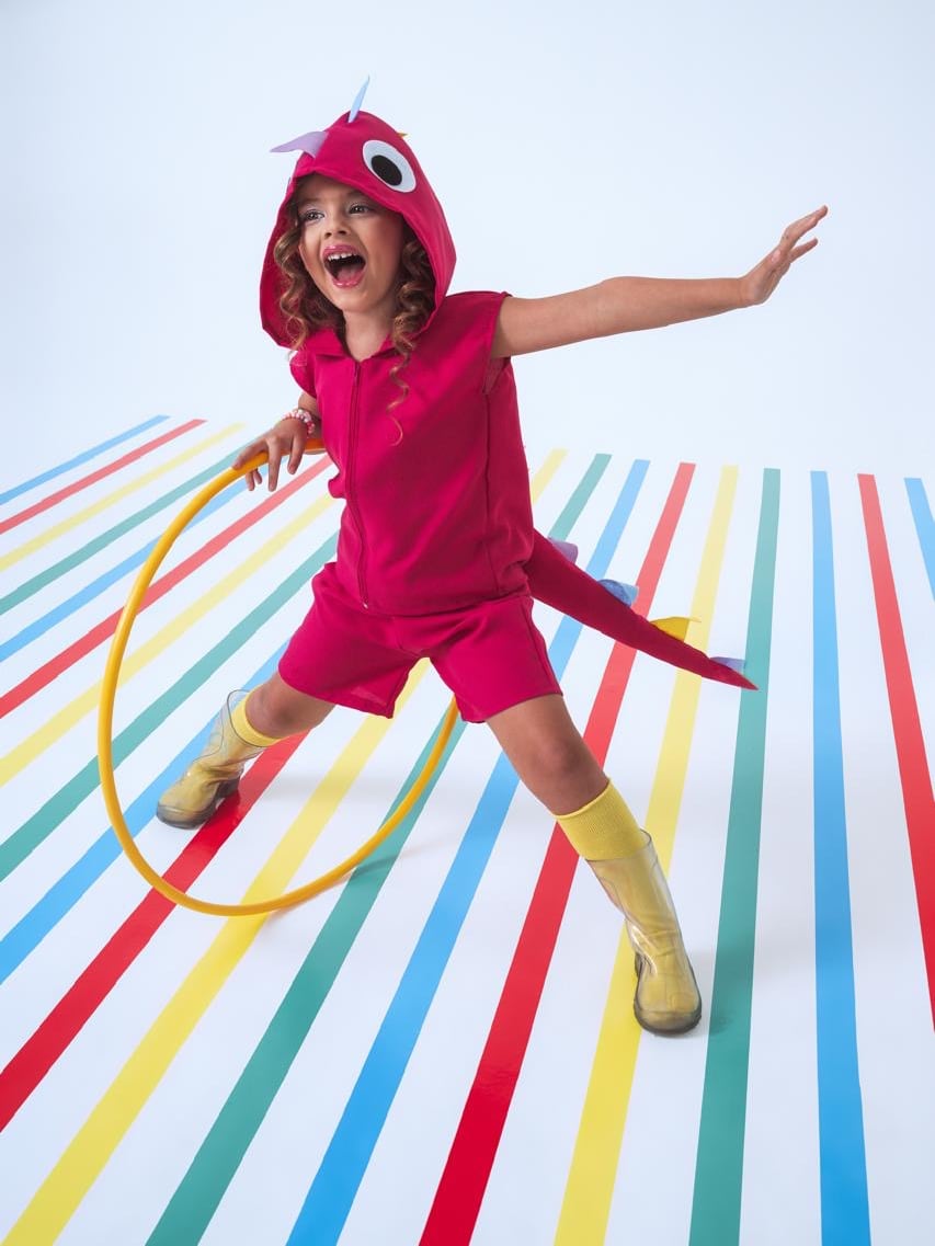 Menina com a fantasia de dinossauro rosa, capuz com olhos e cauda com espinhos, rindo de braços abertos