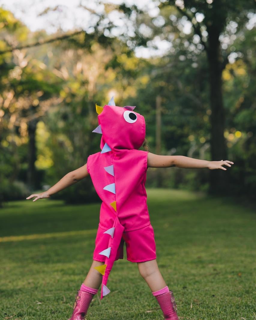Criança de costas num gramado, de braços abertos, com a fantasia de dinossauro rosa: espinhos nas costas e cauda