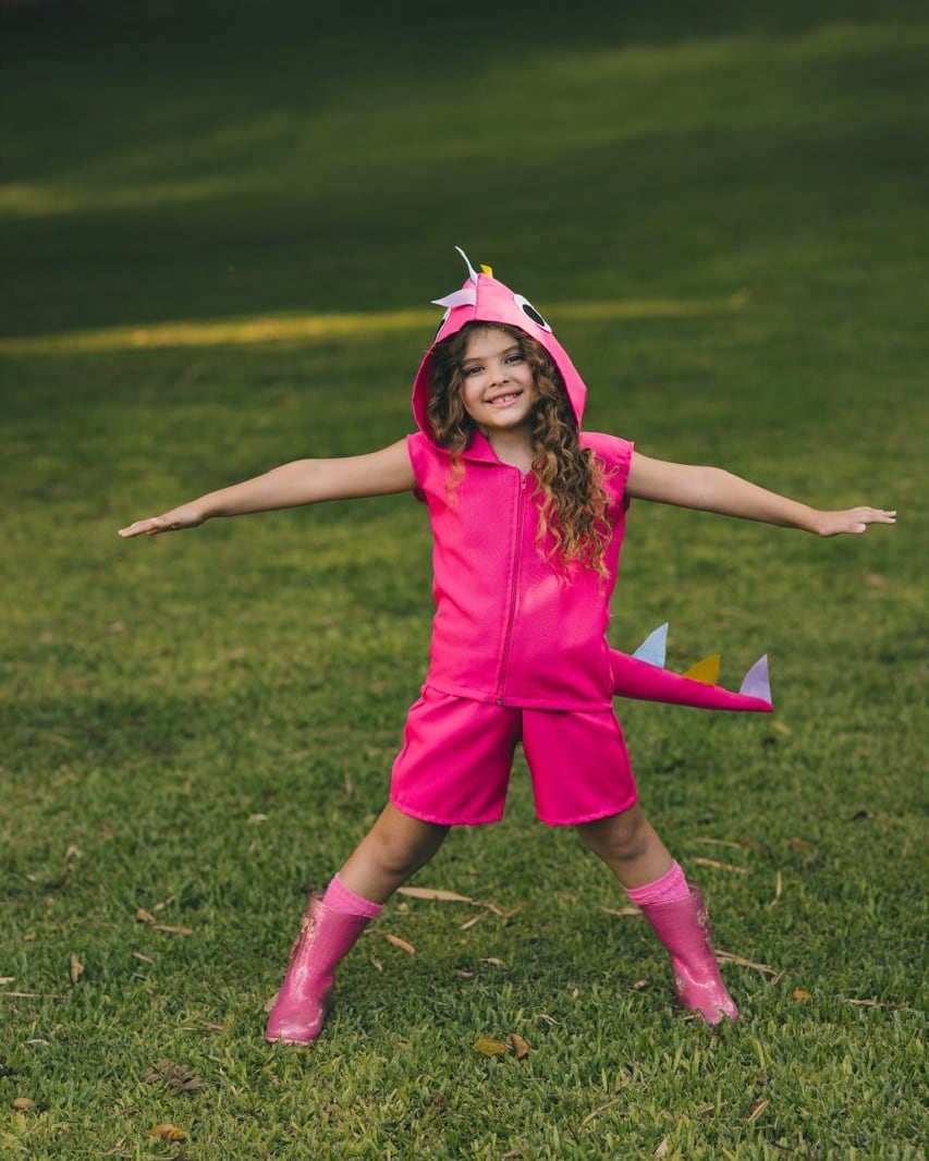 Criança com a fantasia de dinossauro rosa, sorrindo de braços abertos num gramado