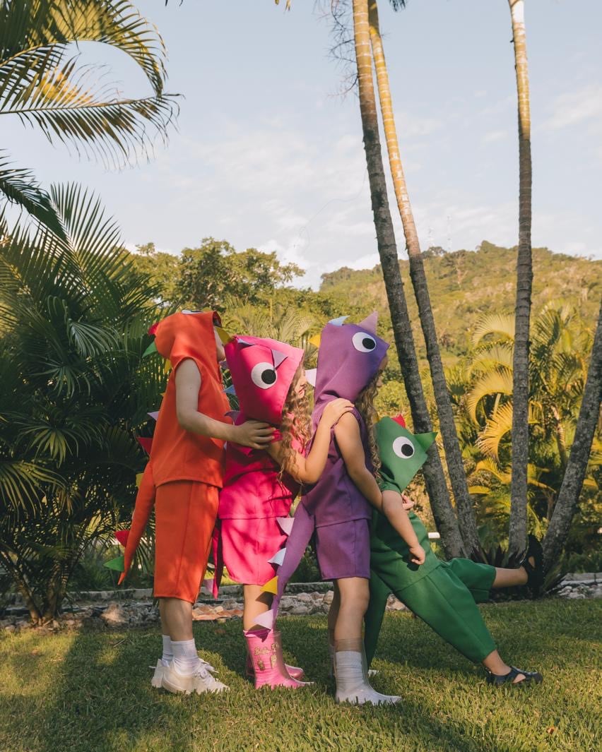 Quatro crianças abraçadas num jardim com palmeiras, com a fantasia de dinossauro nas cores laranja, rosa, roxa e verde