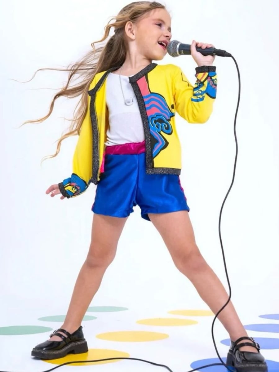 Menina com a fantasia Guerreiros do K-pop, jaqueta amarela estampada e short azul, cantando ao microfone